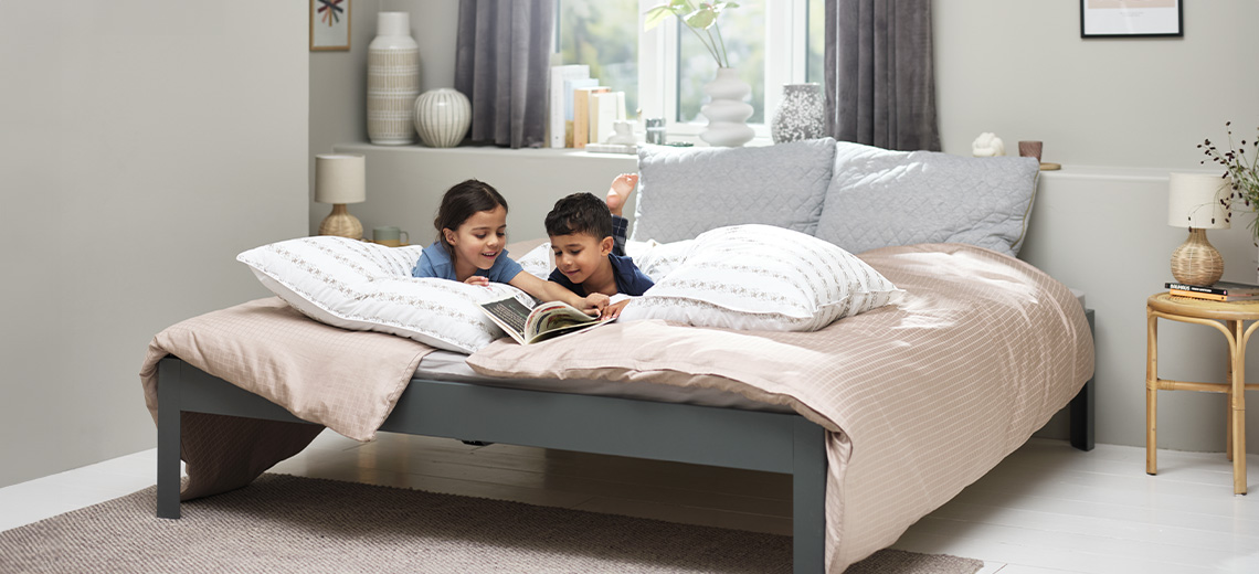 Zwei Kinder lesen ein Buch auf einem Doppelbett mit Bettdecken, Kopfkissen und Zierkissen