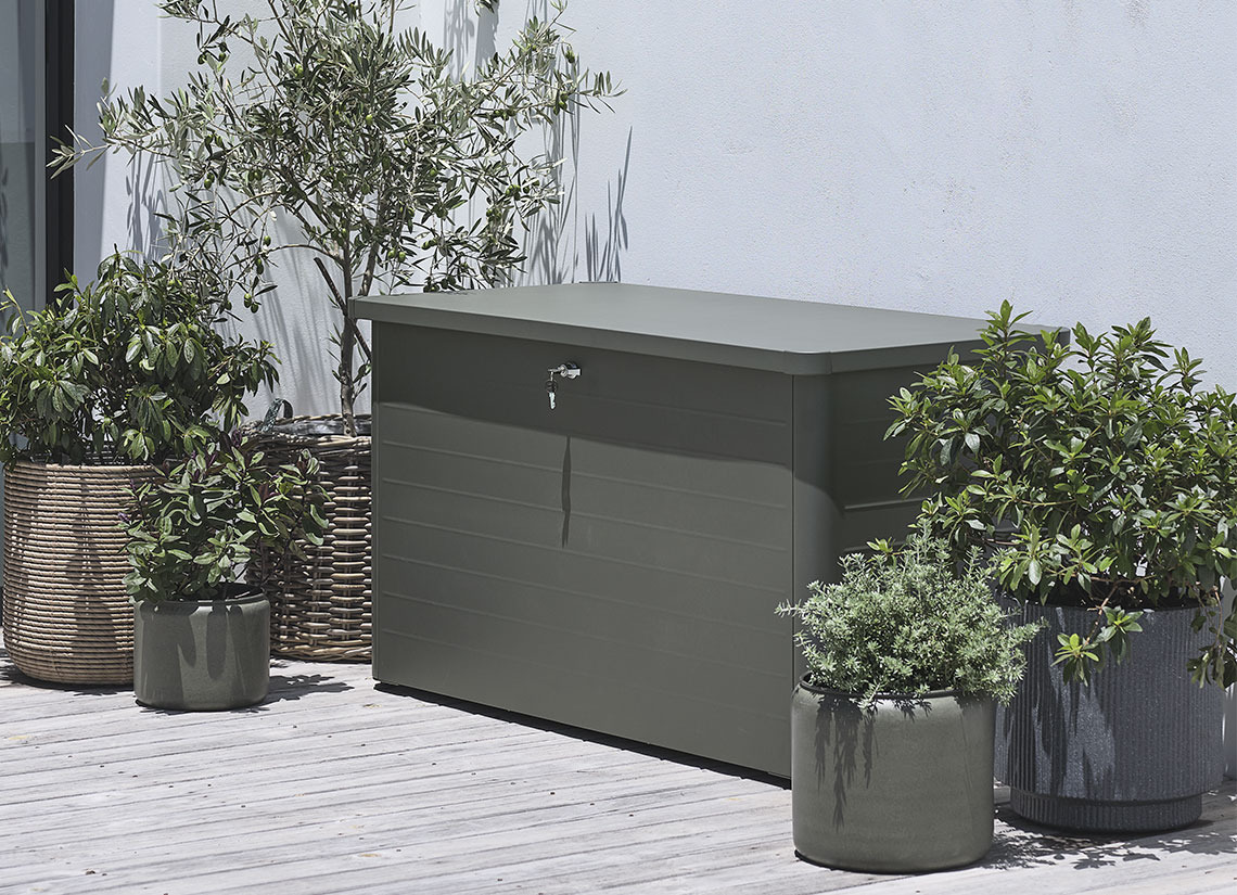 Auflagenbox DREJET ist in zwei verschiedenen Größen erhältlich Graue Gartenbox auf einer Terrasse mit Pflanzen und Pflanzgefäßen