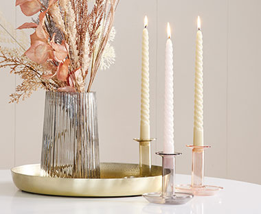 Gedrehte Stabkerzen in Kerzenständern aus Glas mit Tablett, Vase und Trockenblumen auf einem Tisch