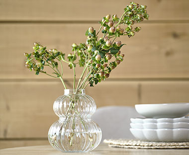 Glasvase mit grünen Blumen neben Schusseln auf einem Tisch vor einer Holzwand